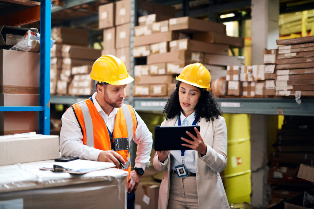 Logistik-Partner schauen in einer Lagerhalle auf ein Tablet und unterhalten sich.